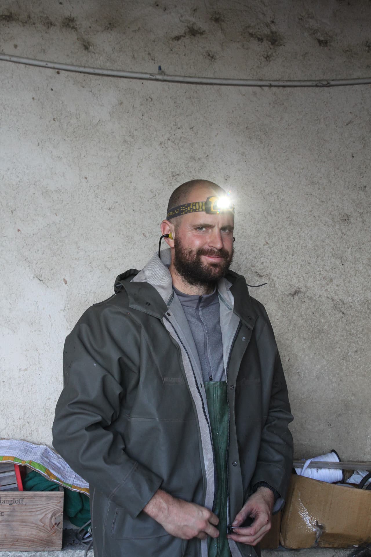 Portrait dans la cave : lampe frontale allumée sur le front, bouchons d'oreilles jaunes, veste de travail olive sur salopette verte, devant un mur de béton clair avec caisses et enrouleurs de corde en arrière-plan